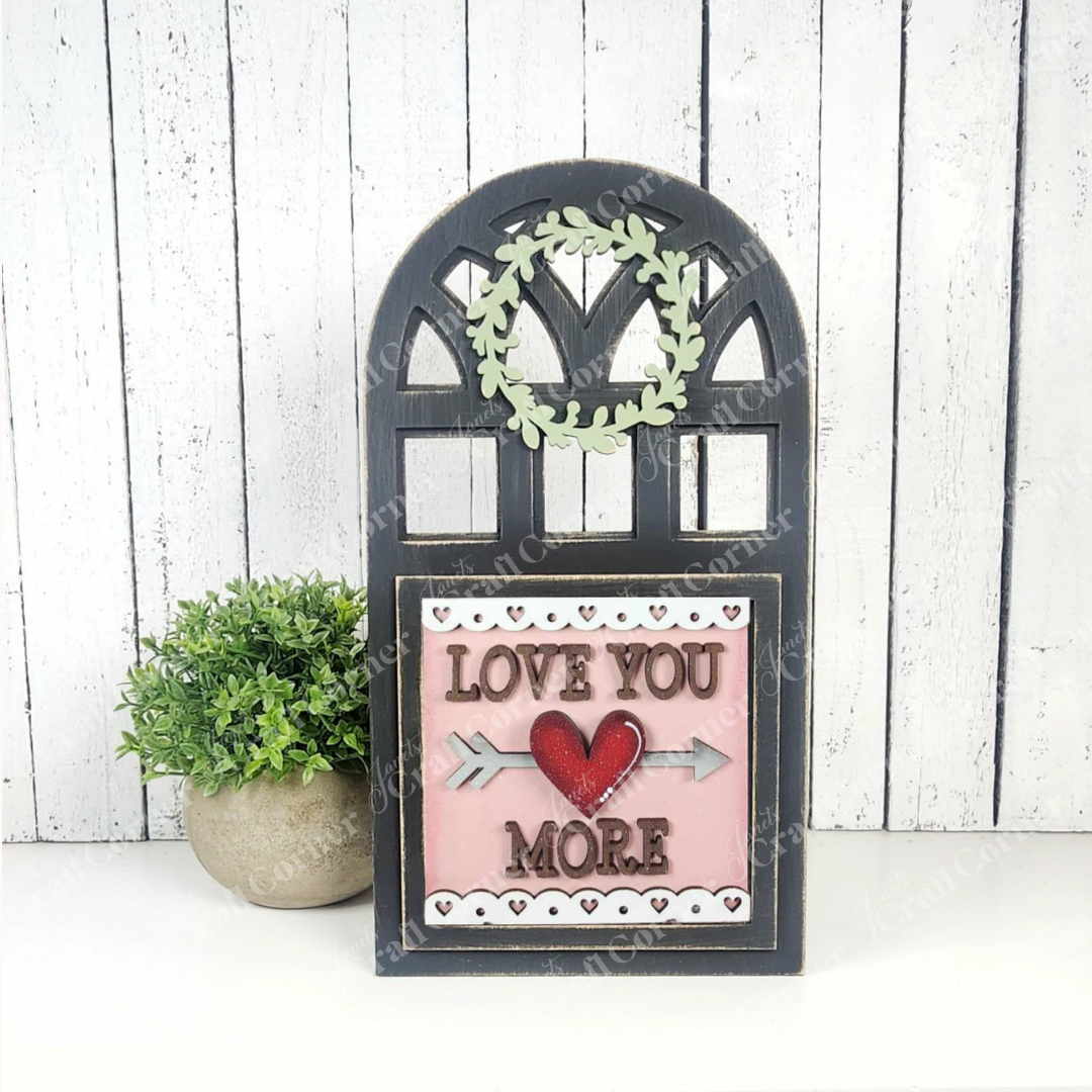 The Arch Window Interchangeable Frame for Squares by Janet's Craft Corner displays a heart and arrow "Love You More" sign beside a green potted plant, bringing charming DIY farmhouse décor to any space.