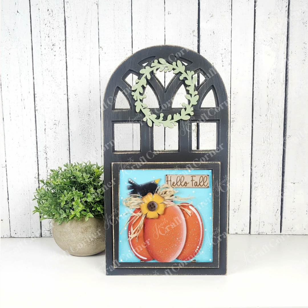 The Arch Window Interchangeable Frame for Squares DIY Farmhouse Decor Kit by Janet's Craft Corner features a black arched frame, leafy wreath, and "Hello Fall" message with painted pumpkins, displayed beside a potted plant on a white wooden background.