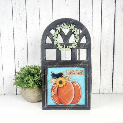 The Arch Window Interchangeable Frame for Squares DIY Farmhouse Decor Kit by Janet's Craft Corner features a black arched frame, leafy wreath, and "Hello Fall" message with painted pumpkins, displayed beside a potted plant on a white wooden background.