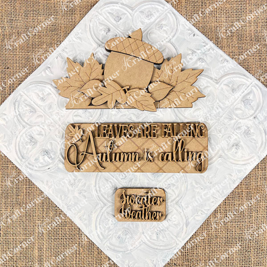 The Autumn Is Calling Interchangeable Insert, a DIY home decor kit from Janet's Craft Corner, showcases hand-painted autumn-themed designs on a textured white and burlap background. The central pieces highlight an acorn surrounded by leaves with the phrase "Leaves are falling, Autumn is calling," accompanied by a smaller piece featuring "Sweater weather." Ideal for interchangeable insert collections or crafting projects.