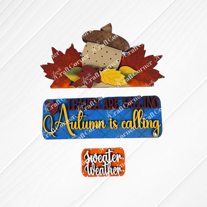 Autumn-themed decorative signs from Janet's Craft Corner, featuring the DIY home decor kit "Autumn Is Calling Interchangeable Insert." The top sign showcases an acorn with leaves and the phrase "Leaves are falling," while the customizable middle sign states, "Autumn is calling," providing an interchangeable touch. The bottom sign reads, "Sweater weather," embellished with leaves.