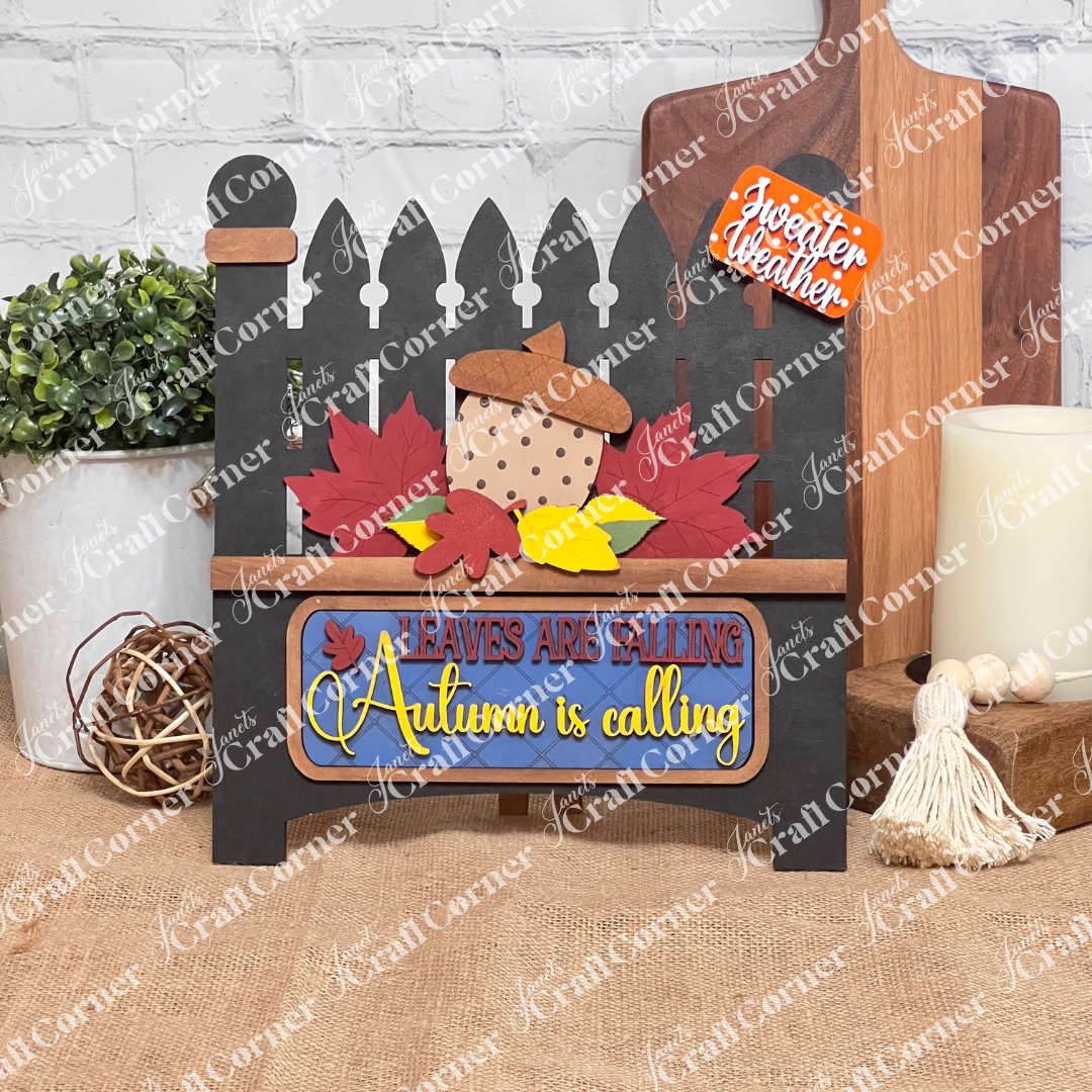 Janet's Craft Corner offers a DIY kit with a white picket fence design, featuring colorful leaves and a large acorn insert. Text reads "Leaves are Falling, Autumn is Calling" with "Sweater Weather" above, perfect for home decor against a charming brick background.