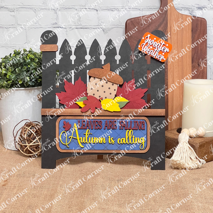 Janet's Craft Corner offers a DIY kit with a white picket fence design, featuring colorful leaves and a large acorn insert. Text reads "Leaves are Falling, Autumn is Calling" with "Sweater Weather" above, perfect for home decor against a charming brick background.