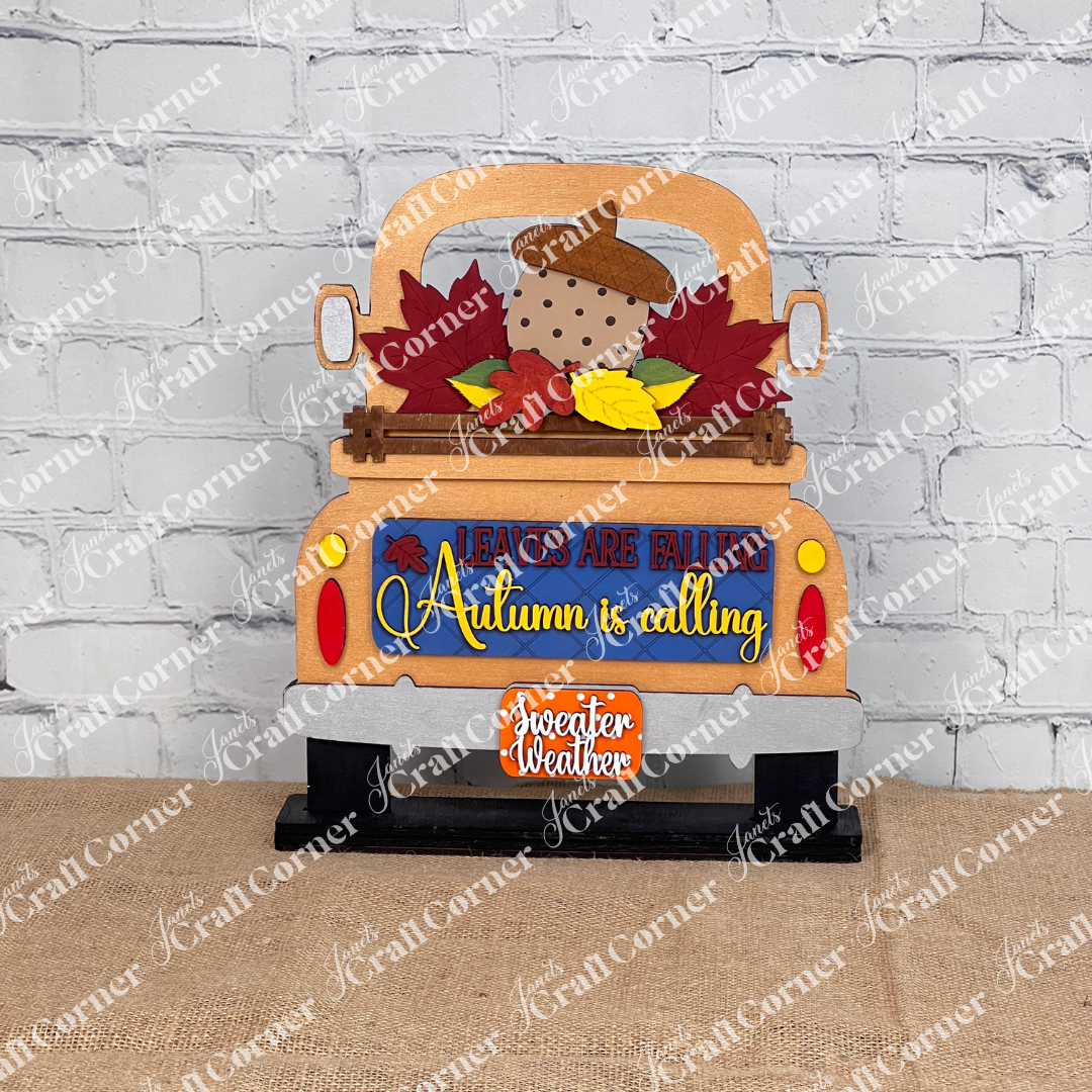 Janet's Craft Corner offers the "Autumn Is Calling Interchangeable Insert," a DIY home decor kit, featuring a decorative piece styled like the back of a copper truck with vibrant leaves and an acorn. The sign displays the message "LEAVES ARE FALLING, Autumn is Calling, sweater weather" and includes an interchangeable insert. It is beautifully set against a brick wall with wooden boards and a candle in the background.