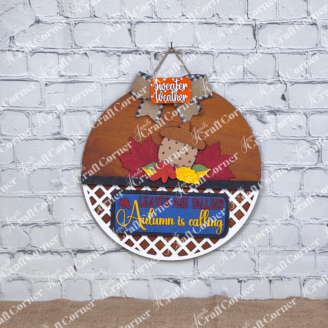 Janet's Craft Corner offers the "Autumn Is Calling Interchangeable Insert," a DIY home decor kit on a door hanger, featuring with vibrant leaves and an acorn. The sign displays the message "LEAVES ARE FALLING, Autumn is Calling, sweater weather" and includes an interchangeable insert. It is beautifully set against a brick wall with wooden boards and a candle in the background.