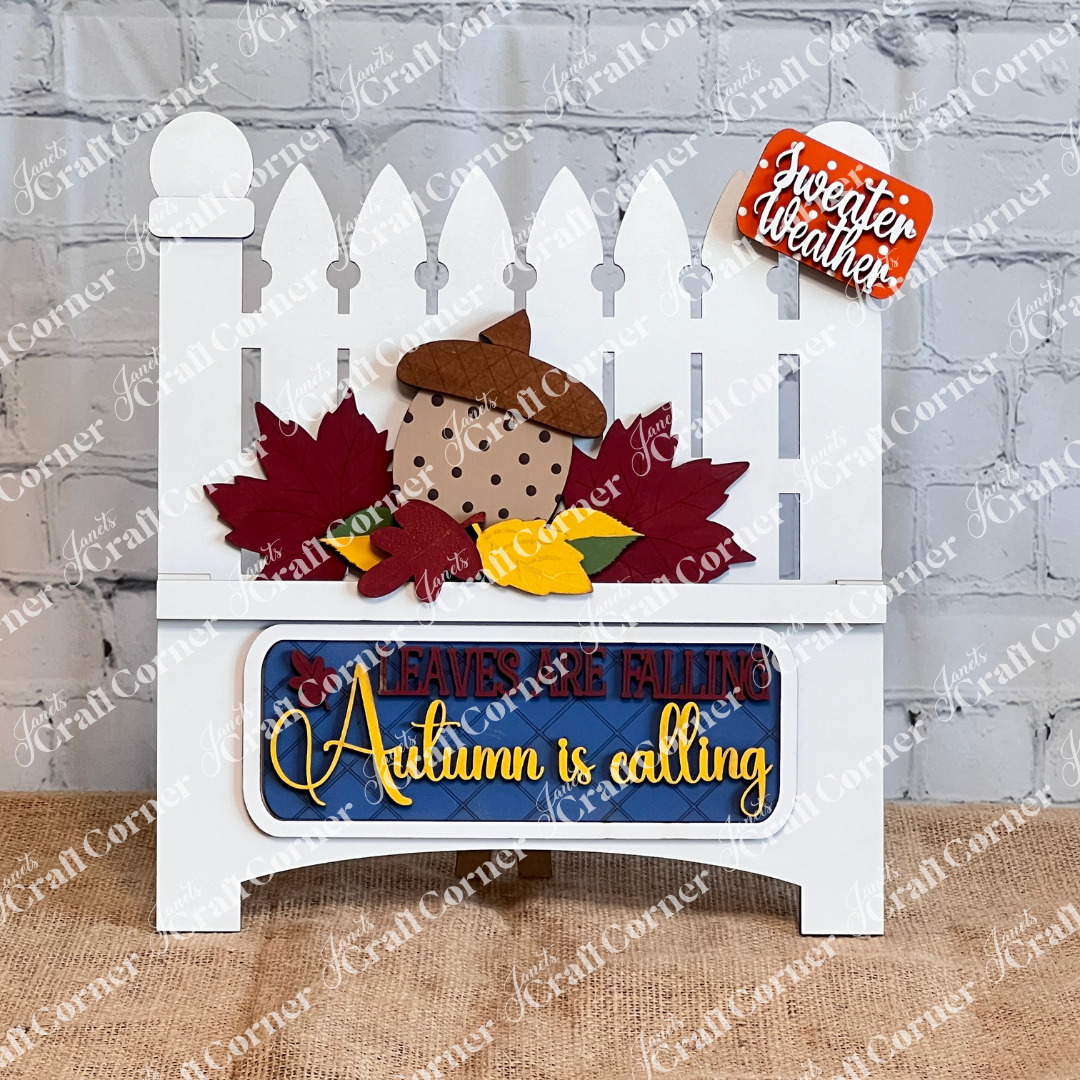 Janet's Craft Corner offers the "Autumn Is Calling Interchangeable Insert," a DIY home decor kit on a white garden gate, featuring with vibrant leaves and an acorn. The sign displays the message "LEAVES ARE FALLING, Autumn is Calling, sweater weather" and includes an interchangeable insert. It is beautifully set against a brick wall with wooden boards and a candle in the background.