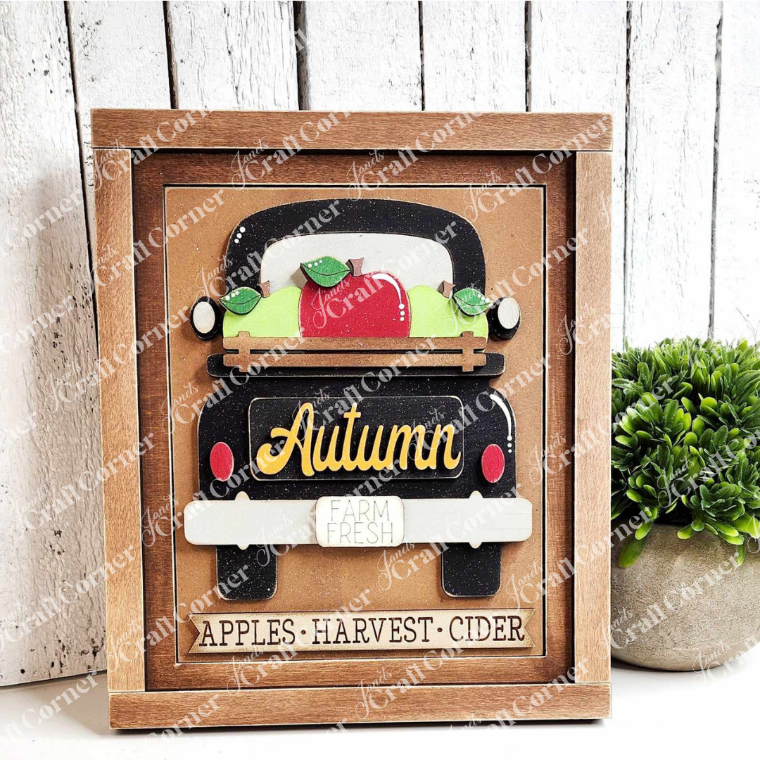 An Autumn Truck Interchangeable Sign from Janet's Craft Corner features a vintage truck overflowing with apples and pumpkins. The words "Autumn" and "Farm Fresh" decorate the truck, while "Apples • Harvest • Cider" is displayed on the frame below. Beside this delightful DIY home decor craft kit, a small plant adds to its charm.