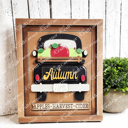 An Autumn Truck Interchangeable Sign from Janet's Craft Corner features a vintage truck overflowing with apples and pumpkins. The words "Autumn" and "Farm Fresh" decorate the truck, while "Apples • Harvest • Cider" is displayed on the frame below. Beside this delightful DIY home decor craft kit, a small plant adds to its charm.