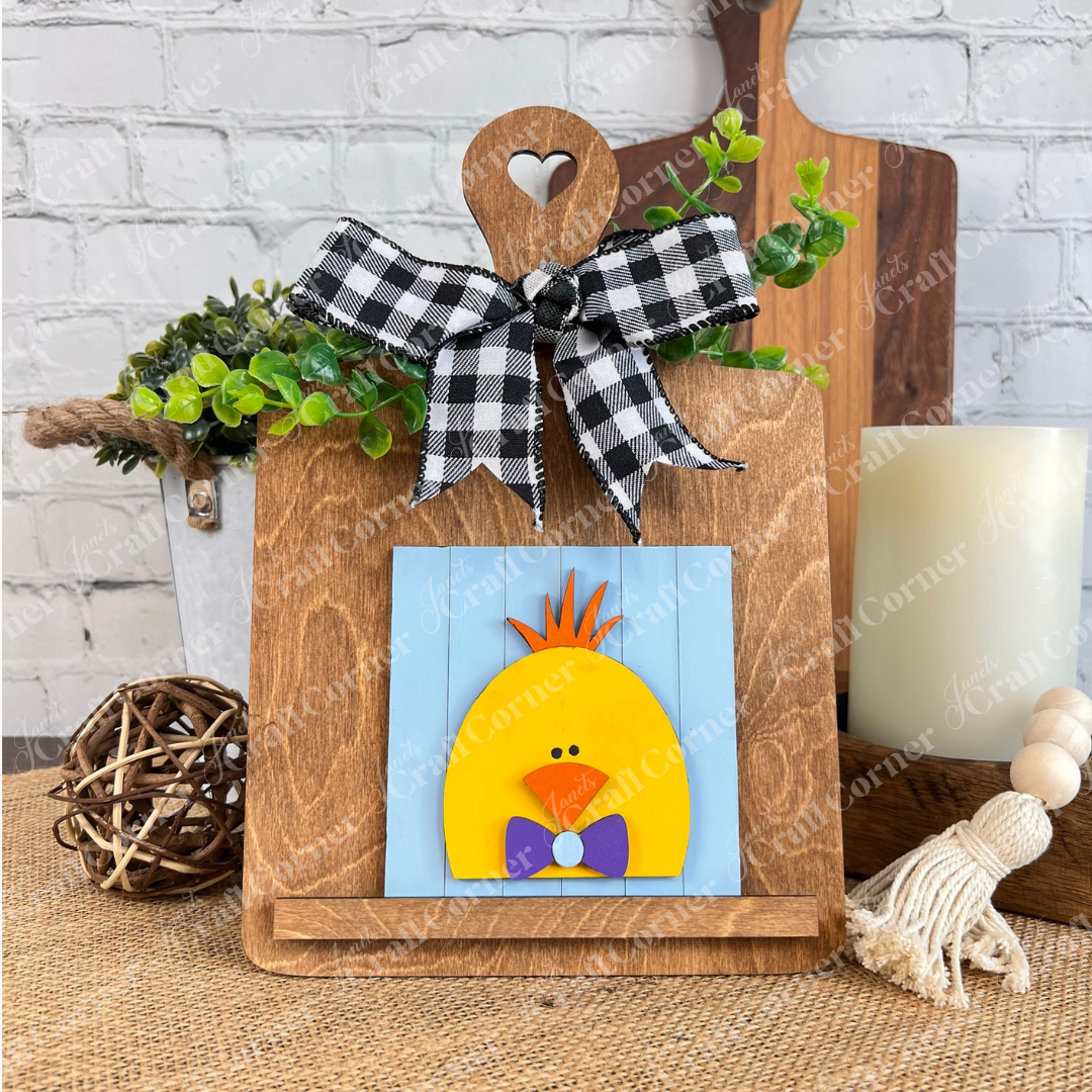 The Hello Easter Squares from Janet's Craft Corner display a yellow chick with an orange tuft and purple bow tie on a blue sign, held by a heart-cutout wooden easel with plaid and greenery, perfect for coffee-themed or rustic DIY decor.