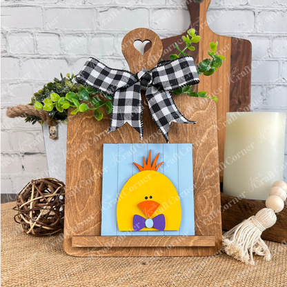 The Hello Easter Squares from Janet's Craft Corner display a yellow chick with an orange tuft and purple bow tie on a blue sign, held by a heart-cutout wooden easel with plaid and greenery, perfect for coffee-themed or rustic DIY decor.