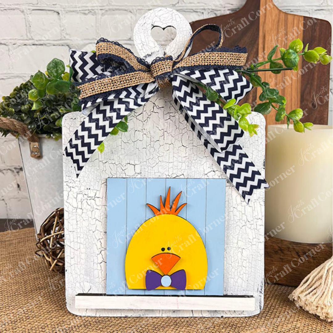 The "Hello Easter Squares" by Janet's Craft Corner is a charming white wooden plaque with a heart cutout, chevron and burlap bows, plus a cute yellow chick—perfect for DIY craft lovers wanting a handmade Easter touch.