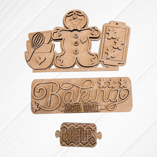 Wooden cookie-themed cutouts are set on a textured surface. The top cutout showcases a gingerbread figure along with baking tools, while the middle bears the phrase "Baking Spirits Bright." This DIY Craft Kit from Janet's Craft Corner is ideal for creating personalized home décor and features the Baking Spirits Bright Interchangeable Insert labeled "Cookies.