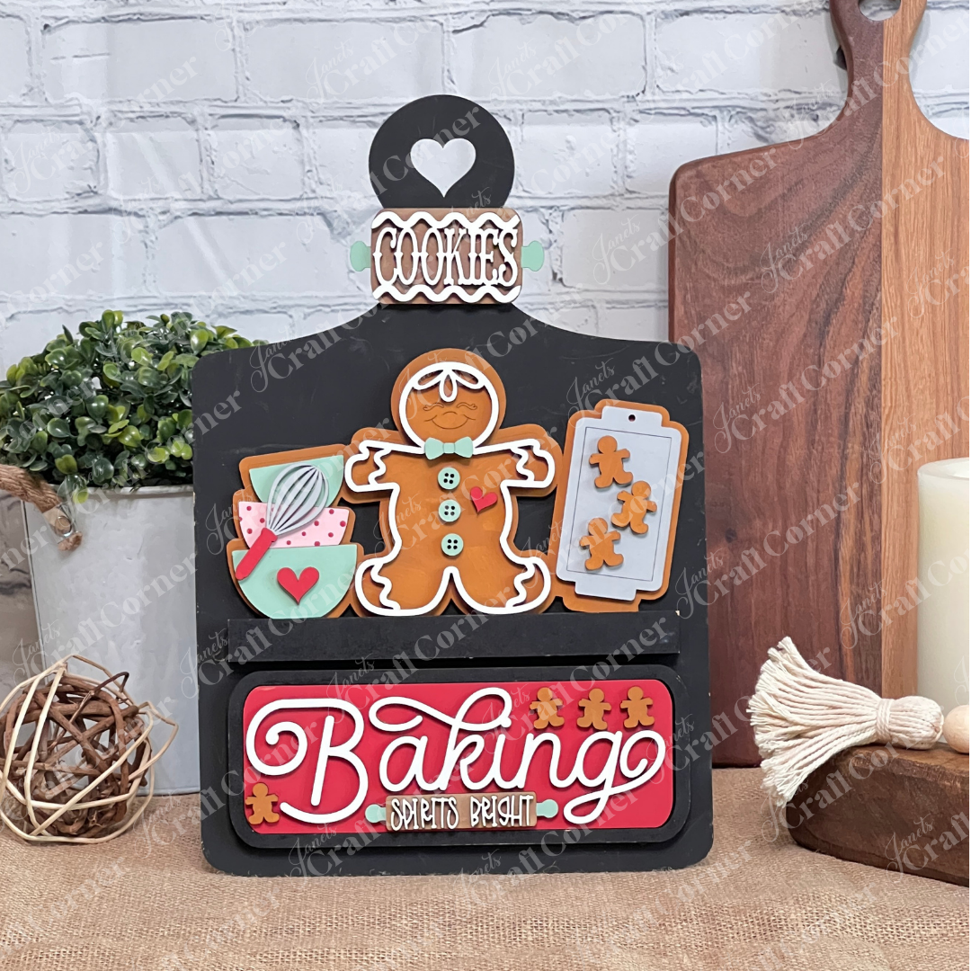 The Breadboard Interchangeable Base with Insert by Janet's Craft Corner features a DIY wooden baking theme. It boasts a gingerbread man, utensils in a bowl, and a cookie tray against a white brick wall. A red "Baking Spirits Bright" sign and heart cutout labeled "Cookies" enrich your home decor.