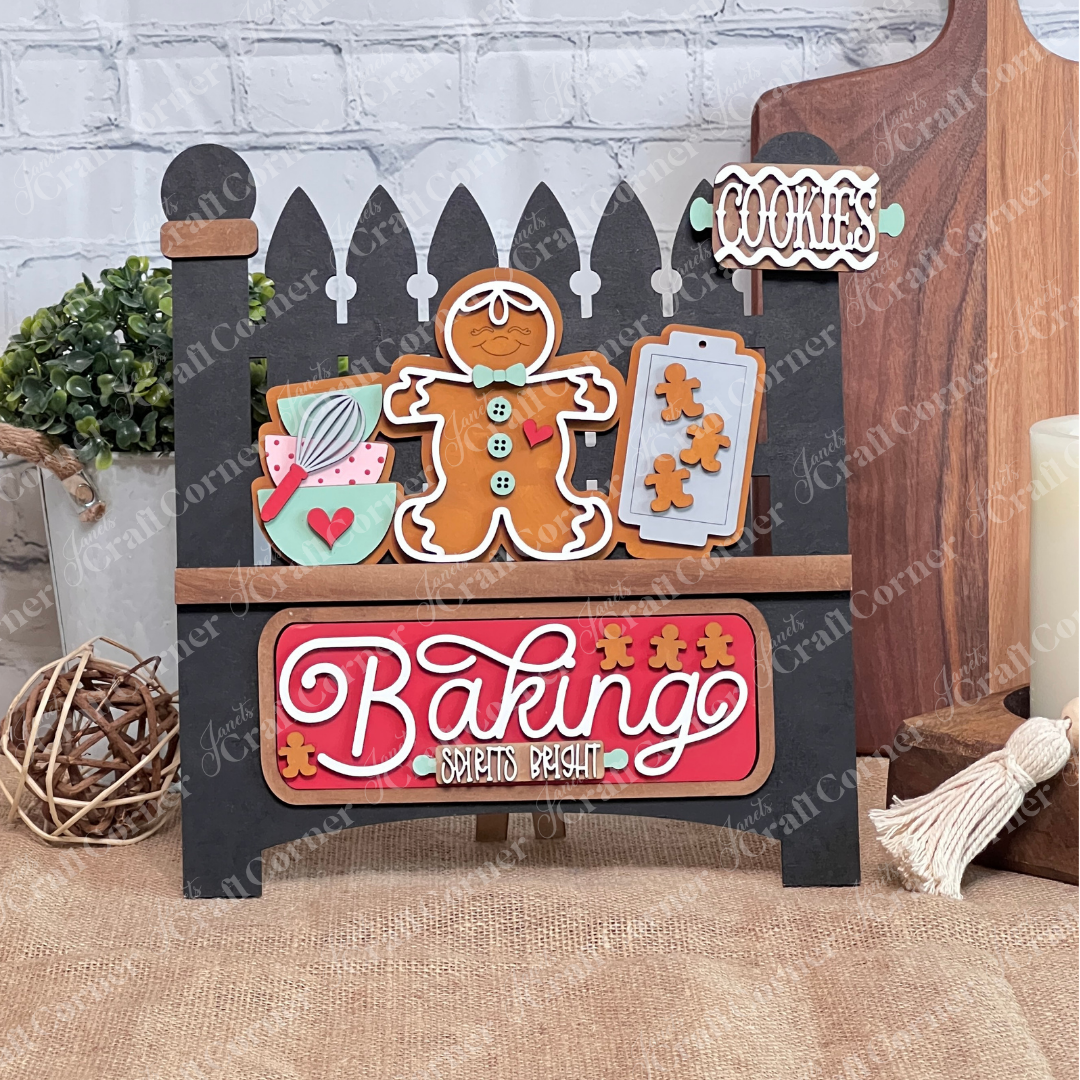 A black garden fence set against a brick wall displays gingerbread-themed decorations, featuring cookie-shaped elements and a "Baking Spirits Bright" interchangeable insert from Janet's Craft Corner. This delightful arrangement, topped with a "Cookies" sign, offers the perfect opportunity for personalization to create an ideal DIY craft kit experience.