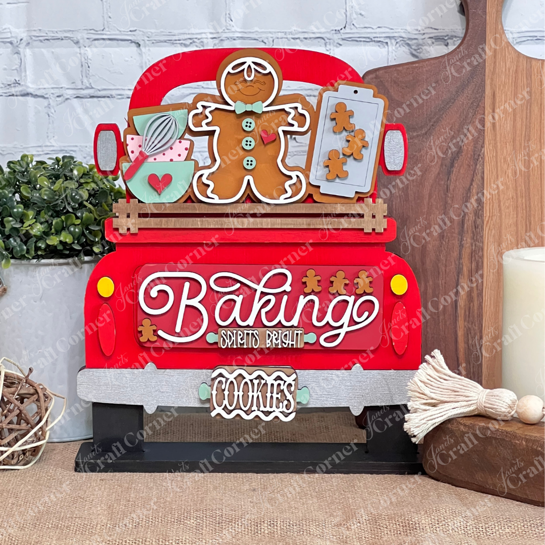 The Antique Truck Base with Insert DIY kit by Janet's Craft Corner features a wooden decoration with baking-themed items like a gingerbread man, cookie sheet, and whisk. It displays "Baking Spirits Bright" and "Cookies" against a white brick wall background, perfect for home décor enthusiasts!.