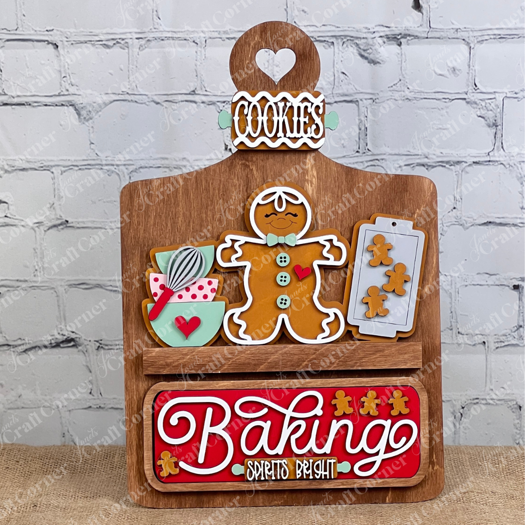 A wooden stand set against a brick wall displays gingerbread-themed decorations, featuring cookie-shaped elements and a "Baking Spirits Bright" interchangeable insert from Janet's Craft Corner. This delightful arrangement, topped with a "Cookies" sign, offers the perfect opportunity for personalization to create an ideal DIY craft kit experience.