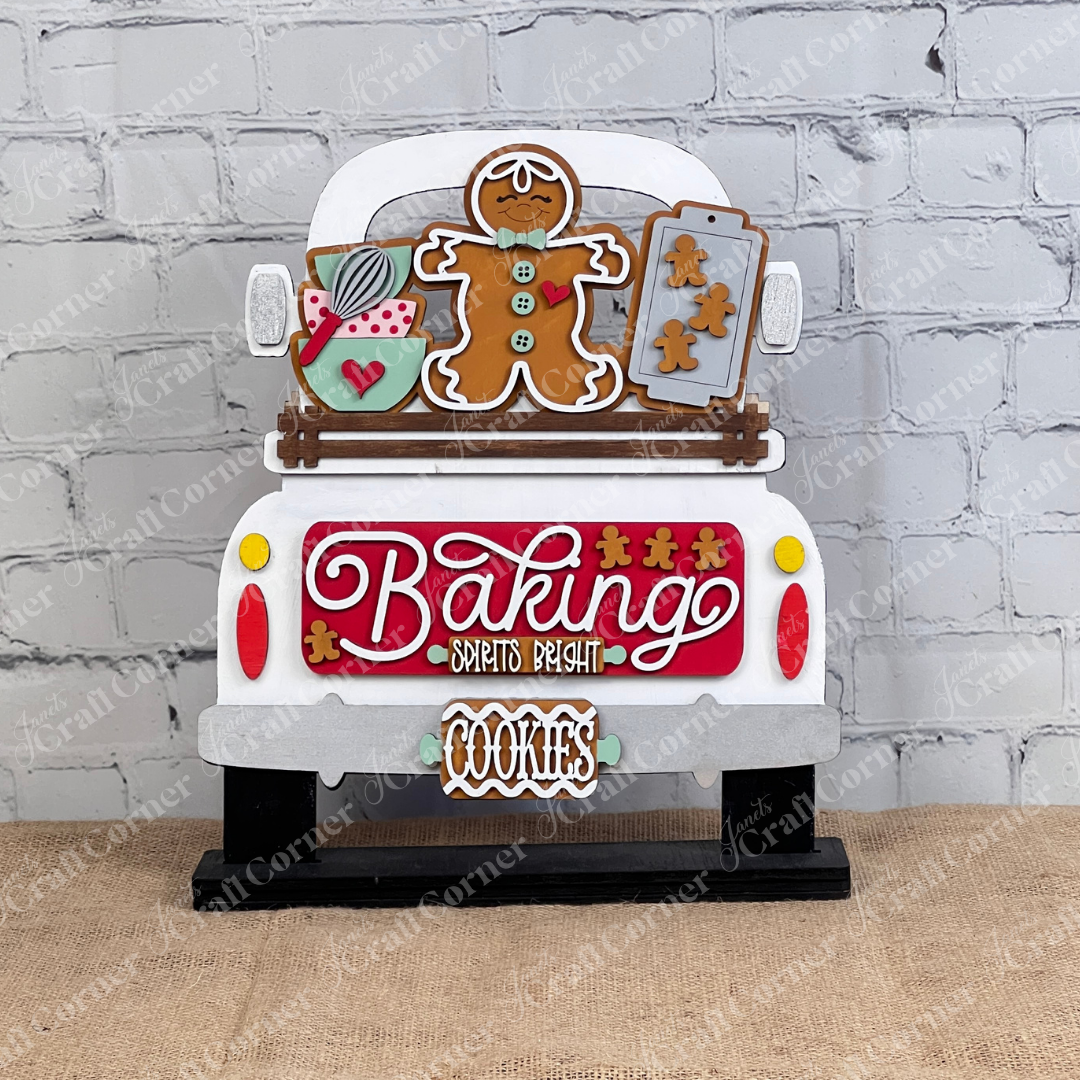 The "Baking Spirits Bright Interchangeable Insert - DIY Craft Kit" by Janet's Craft Corner is a charming piece shaped like the back of a truck, featuring festive elements such as a gingerbread man, cookie sheets, and a mug. The sign displays the word "Baking" prominently with "Spirits Bright" in smaller text and includes a hanging plaque reading "COOKIES." It's perfect for personalized home décor, showcased beautifully against a white brick wall.
