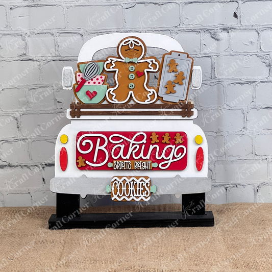 The "Baking Spirits Bright Interchangeable Insert - DIY Craft Kit" by Janet's Craft Corner is a charming piece shaped like the back of a truck, featuring festive elements such as a gingerbread man, cookie sheets, and a mug. The sign displays the word "Baking" prominently with "Spirits Bright" in smaller text and includes a hanging plaque reading "COOKIES." It's perfect for personalized home décor, showcased beautifully against a white brick wall.