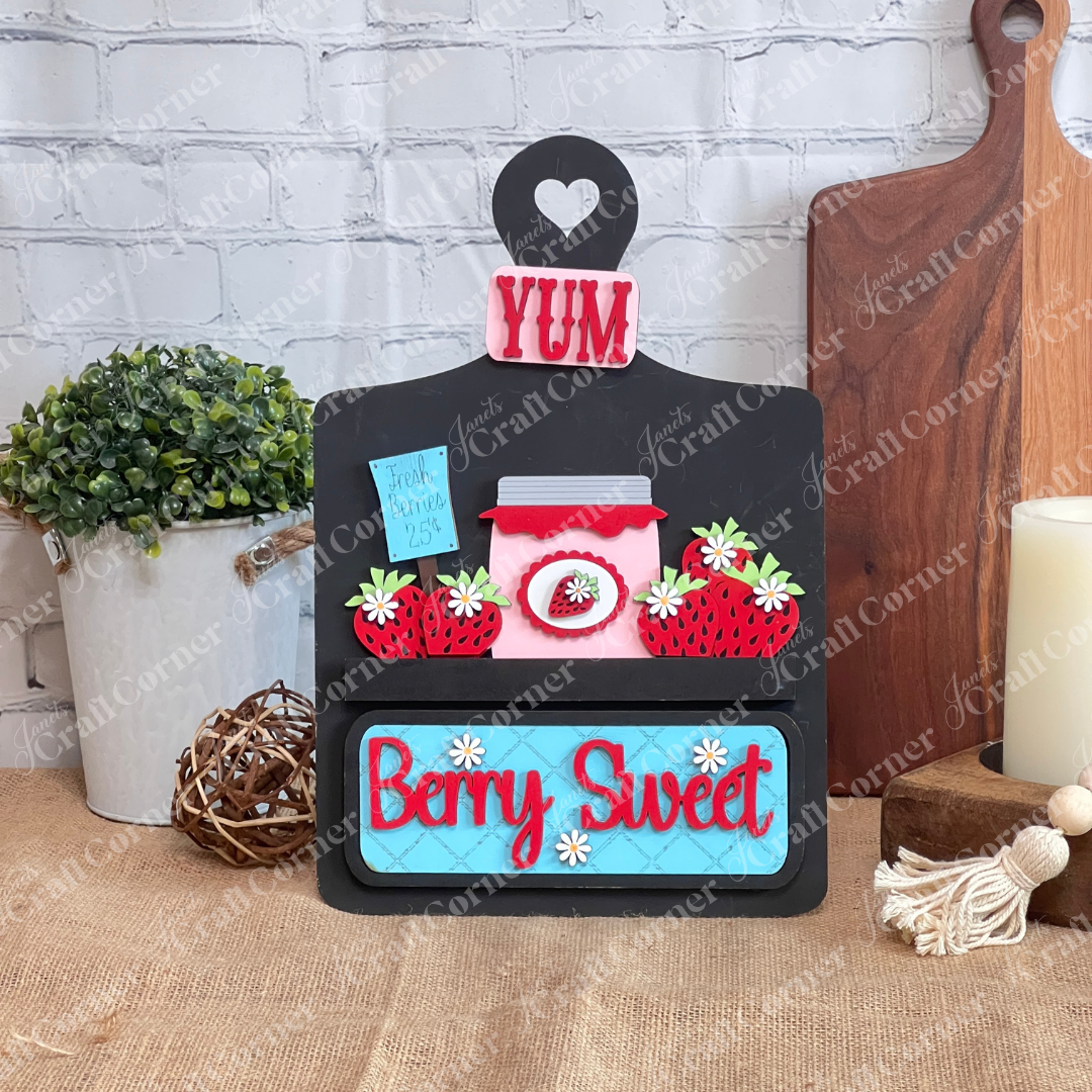 The Berry Sweet Interchangeable Insert from Janet's Craft Corner is a DIY seasonal home decor craft kit featuring a wooden board painted black with "YUM" in vinyl, a jar with strawberries, and a blue "Fresh Berries 25¢" sign against a white brick wall.