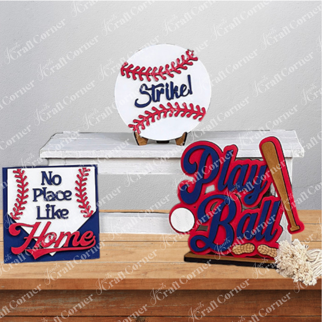A wooden chair displays a Baseball Mini Shelf Sitters from Janet's Craft Corner, featuring a baseball-themed home décor with a DIY kit charm. Signs reading "Strike!", "No Place Like Home," and "Play Ball" enhance your shelves, evoking the essence of a delightful baseball game.