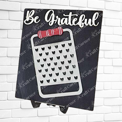 Janet's Craft Corner Kitchen Switchable Farmhouse Sign features "Be Grateful" above a cheese grater with red handle on a dark background—a DIY home decor craft kit, perfect rustic touch for your kitchen, displayed on a white brick wall.
