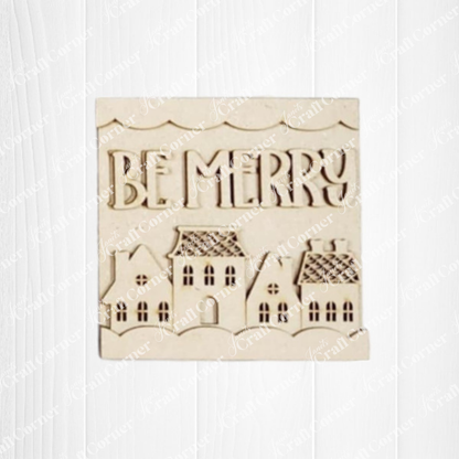 A cream-colored plaque reading "BE MERRY" sits above embossed houses on a wood background—perfect holiday décor or pairing with Janet's Craft Corner Gingerbread Mini Shelf Sitters DIY craft kit.
