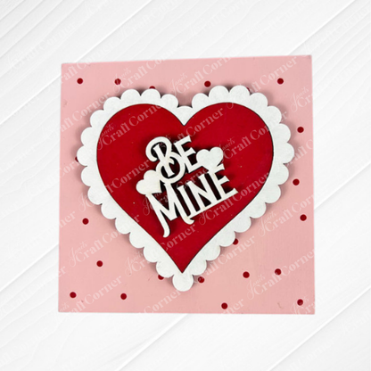 The Valentine Squares by Janet's Craft Corner is a square pink card with red polka dots and a scalloped red-and-white heart in the center. "Be Mine" is written inside the cheerful heart, perfect for DIY seasonal home or tiered tray decor.