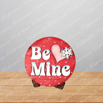 The Valentine Snowmen Decor Set from Janet's Craft Corner features a round red sign with "Be Mine" in white text, a pink heart, and white snowflake—perfect with snowman wood cut-outs on a wooden stand for festive DIY decorating.