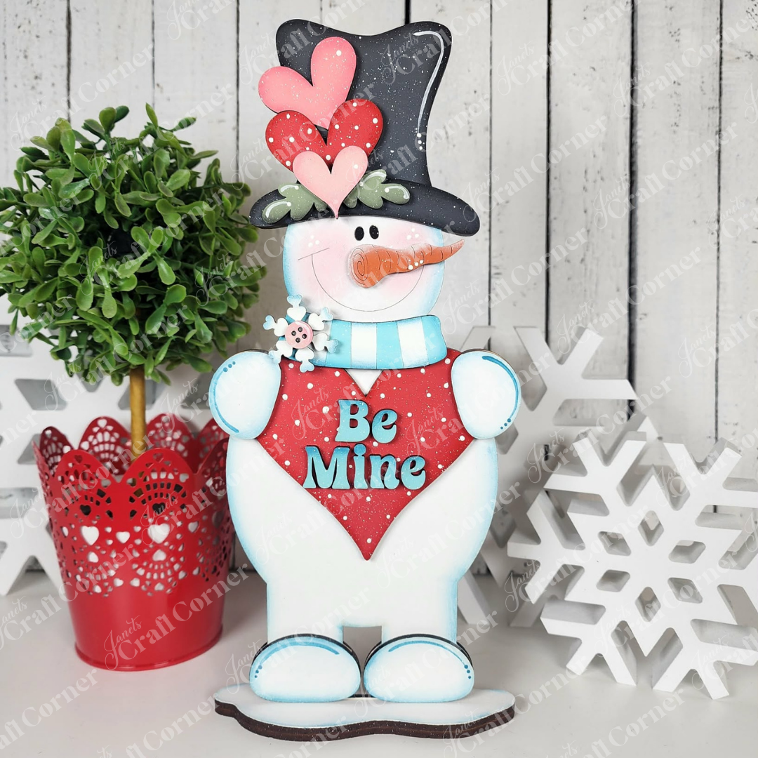 The Valentine Snowmen Decor Set from Janet's Craft Corner includes a cheerful snowman shelf sitter with a top hat, scarf, and red "Be Mine" heart, plus wood snowflake cut-outs and a small green potted plant for festive DIY home decor.