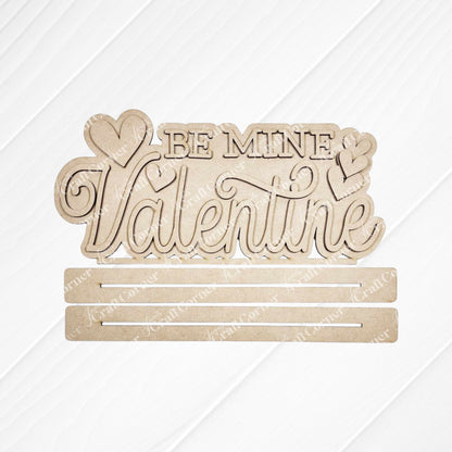 Add charm to your home décor with the "Be Mine Valentine" Word Art from Janet's Craft Corner. This hand-painted wooden cutout sign features cursive text and heart shapes on a white background, complete with two horizontal slots for DIY projects.