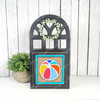 Janet's Craft Corner's Summer Beach Squares DIY kit includes a black wooden frame with a wreath and beach ball design on a blue square, displayed beside a green potted plant against a white wood wall—perfect for seasonal home decor.