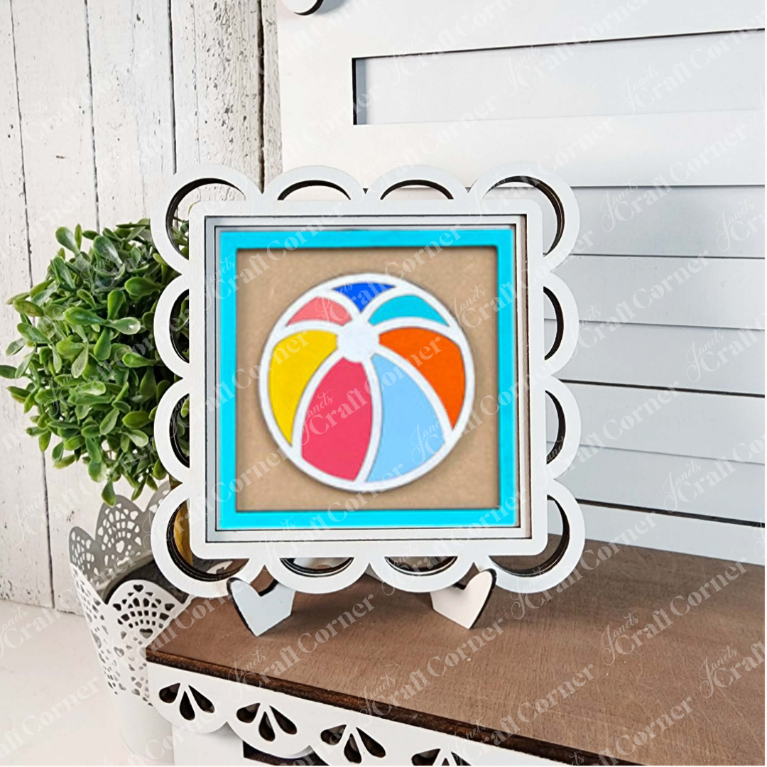 Janet’s Craft Corner’s Summer Beach Squares DIY craft kit features a framed beach ball illustration on a stand, displayed with tiered tray signs and accents, creating cheerful seasonal decor against a white wooden background.