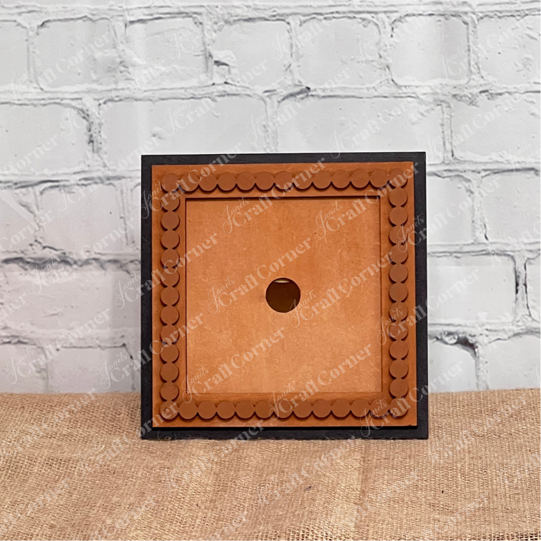 A Heartland Bead Trim Swappable Squares Frame, square and brown with a scalloped border, is shown from the back displaying a central round mounting hole. It rests on textured beige fabric against a white brick wall background.