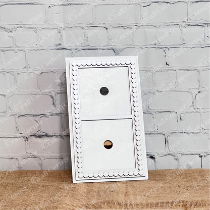 The Heartland Bead Trim Swappable Squares Frame is a white rectangular wooden frame with two cut-out circles and decorative scallop edging. Ideal for DIY craft kits, it stands upright against a white brick wall on burlap.