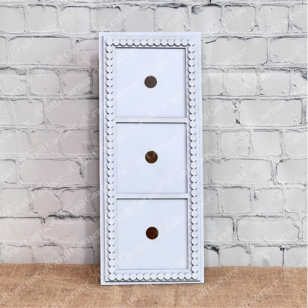 The Heartland Bead Trim Swappable Squares Frame is a white, rectangular wooden organizer with three drawers featuring round hole handles and decorative beading, ideal for storing DIY craft kits against a white brick wall.