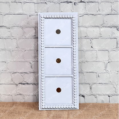 The Heartland Bead Trim Swappable Squares Frame is a white, rectangular wooden organizer with three drawers featuring round hole handles and decorative beading, ideal for storing DIY craft kits against a white brick wall.