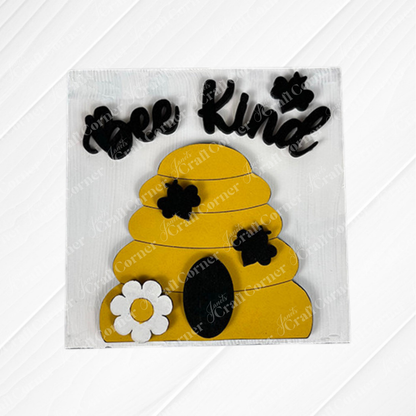 The Bee Swappable Squares from Janet's Craft Corner feature a yellow beehive, black bees, a white flower, and "Bee Kind" on a textured white background—adding handmade charm to your home decor.