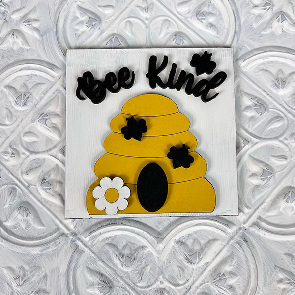 The Bee Swappable Squares from Janet's Craft Corner feature a yellow beehive, black bees, a white flower, and "Bee Kind" on a textured white background—adding handmade charm to your home decor.