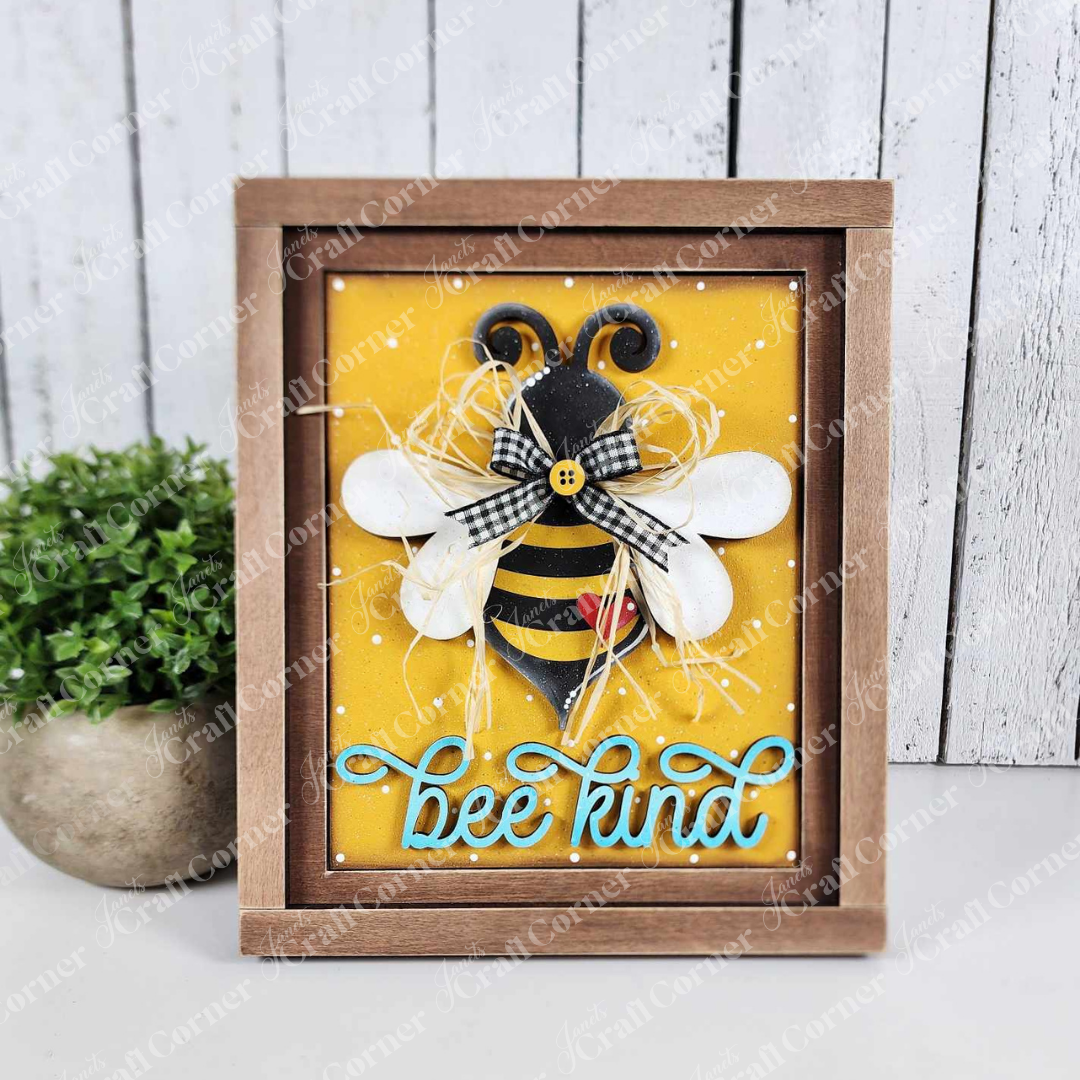 Janet's Craft Corner offers a Bee Kind Farmhouse Sign DIY kit. This painted sample features bee-shaped decor on a yellow background with a gingham bow and blue "Bee Kind" script. It is part of the seasonal home decor series, enhanced by the subtle touch of greenery.