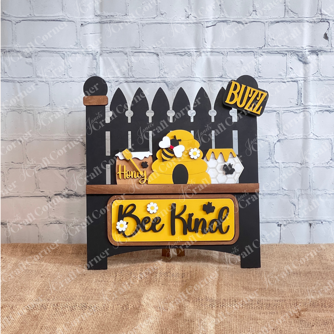 A decorative sign featuring the Bee Interchangeable Insert from Janet's Craft Corner presents a hand-painted black garden fence with a beehive and bees. The piece, which prominently displays "Bee Kind" and a "BUZZ" license plate, stands out against a white brick wall.