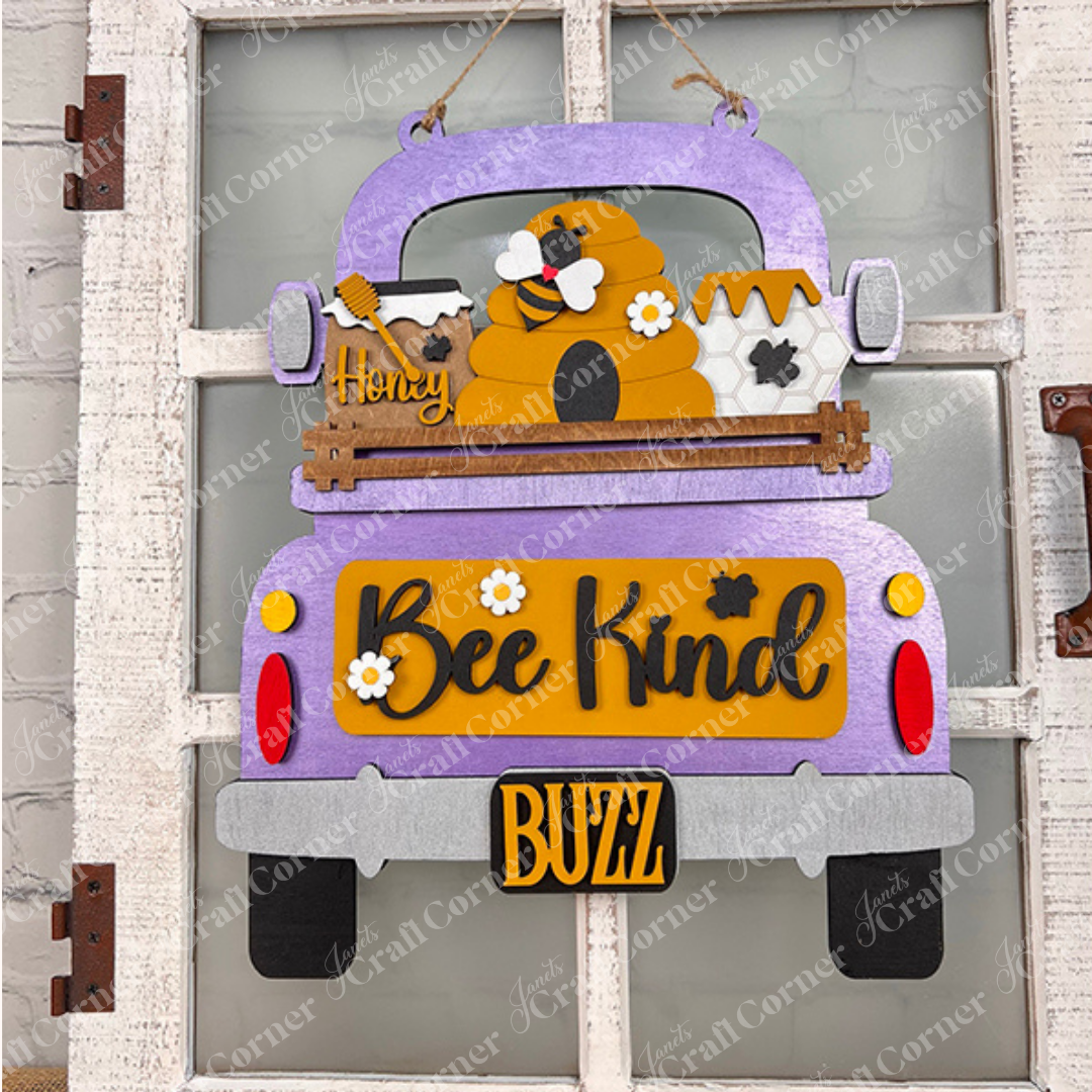 A decorative sign featuring the Bee Interchangeable Insert from Janet's Craft Corner presents a hand-painted purple truck with a beehive and bees. The piece, which prominently displays "Bee Kind" and a "BUZZ" license plate, stands out against a textured gray wall and is enhanced by the brown handle on its white wooden frame.