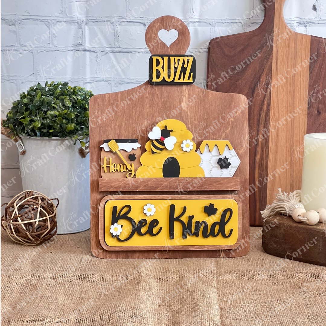 The "Bee Kind" Interchangeable Insert by Janet's Craft Corner features a beehive, honey jars, bee motifs, a "Buzz" label, and plaid bow on a stained breadboard on a burlap base against a white brick wall—perfect for decorating any space with charm.