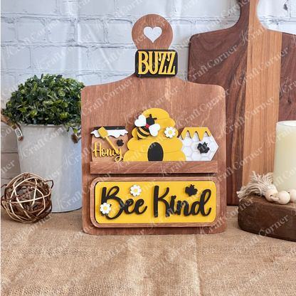 The "Bee Kind" Interchangeable Insert by Janet's Craft Corner features a beehive, honey jars, bee motifs, a "Buzz" label, and plaid bow on a stained breadboard on a burlap base against a white brick wall—perfect for decorating any space with charm.