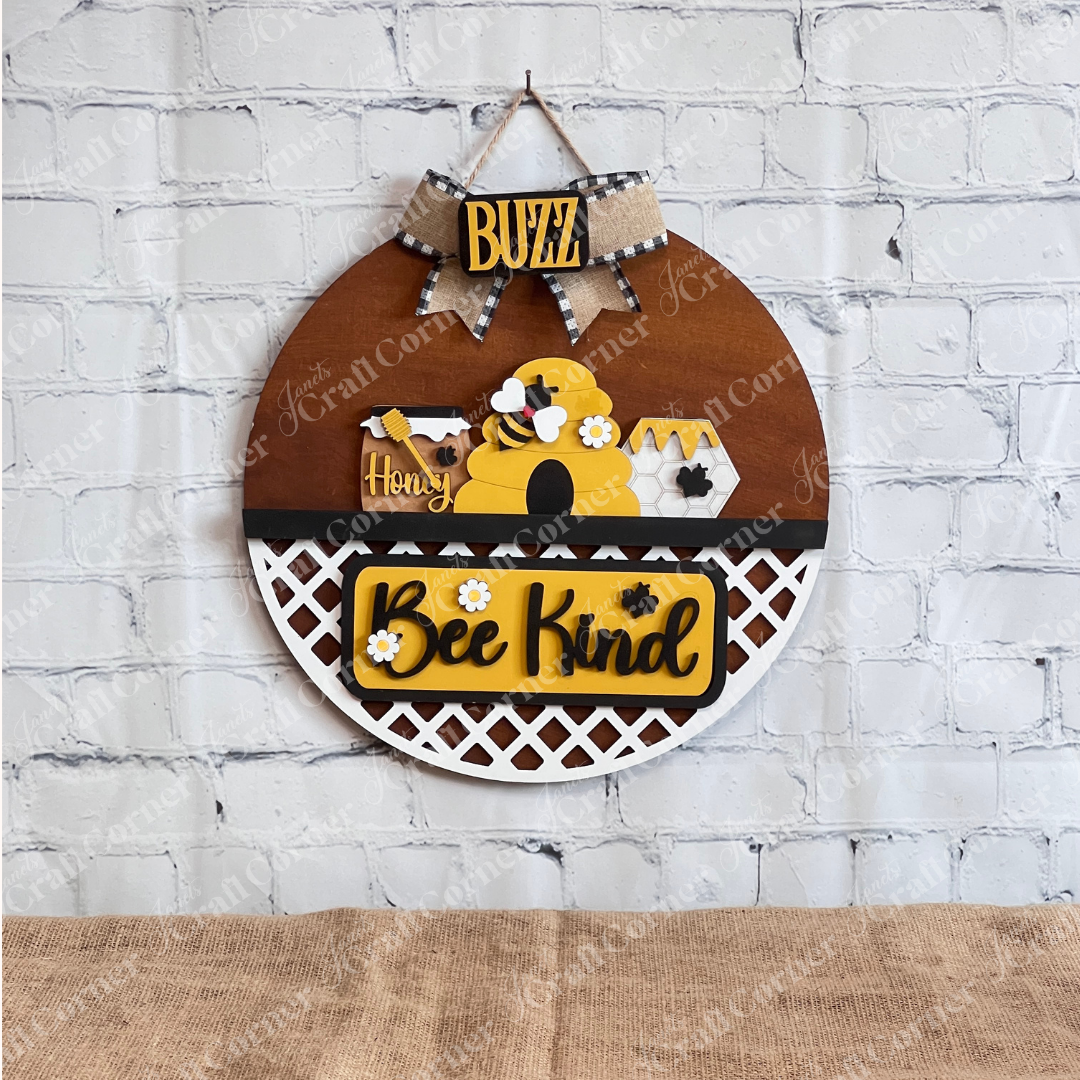 Discover the Bee Interchangeable Insert - Painted from Janet's Craft Corner. This round wooden decorative hanging sign features a charming beehive, bees, and honeycomb motif. It includes the words "Buzz," "Honey," and "Bee Kind" with a white lattice pattern at the bottom, offering versatile decor options with its interchangeable bee inserts.