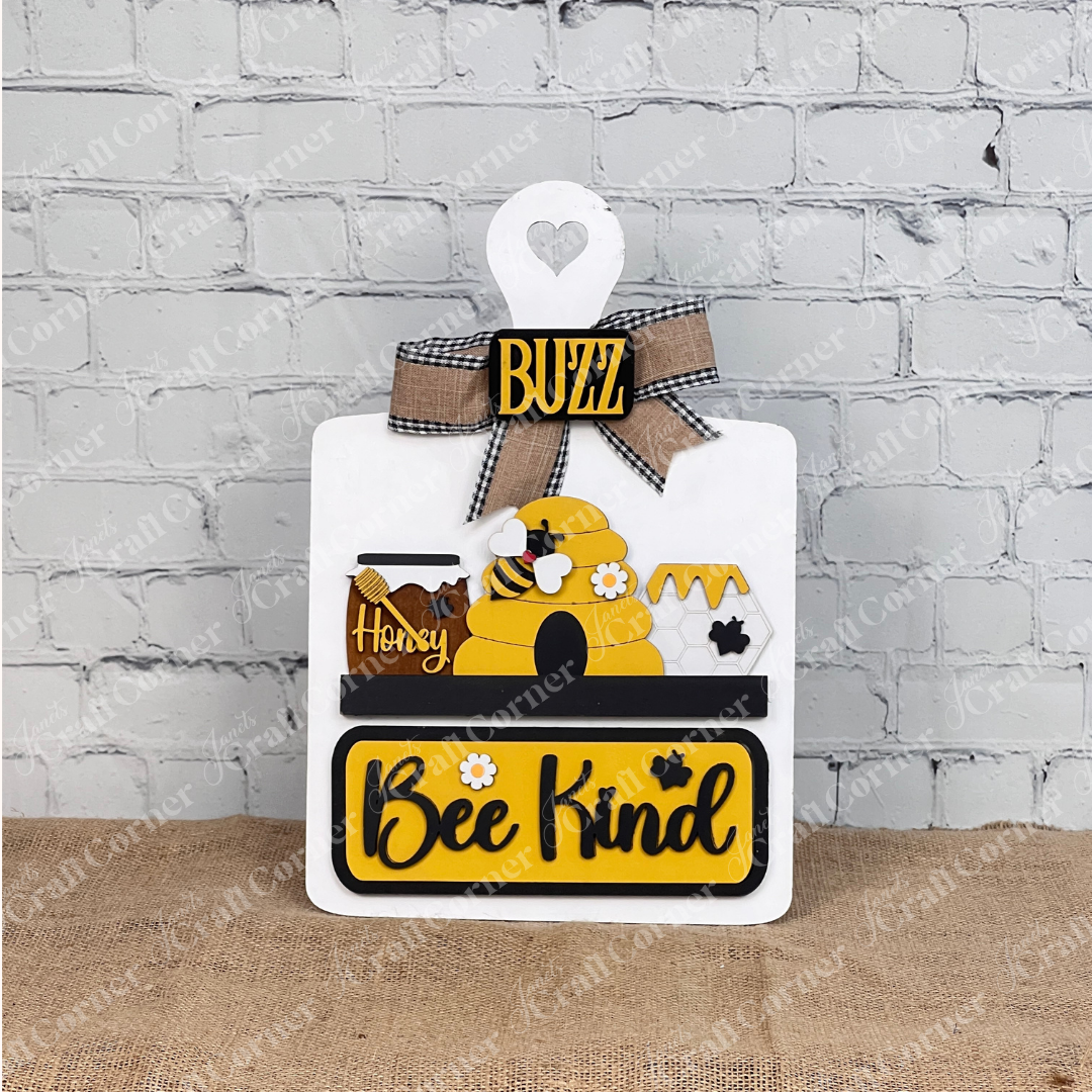 The "Bee Kind" Interchangeable Insert by Janet's Craft Corner features a beehive, honey jars, bee motifs, a "Buzz" label, and plaid bow on a burlap base against a white brick wall—perfect for decorating any space with charm.