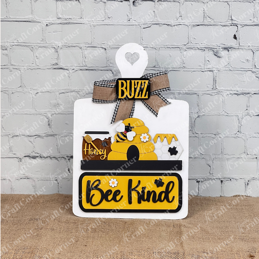 The "Bee Kind" Interchangeable Insert by Janet's Craft Corner features a beehive, honey jars, bee motifs, a "Buzz" label, and plaid bow on a burlap base against a white brick wall—perfect for decorating any space with charm.