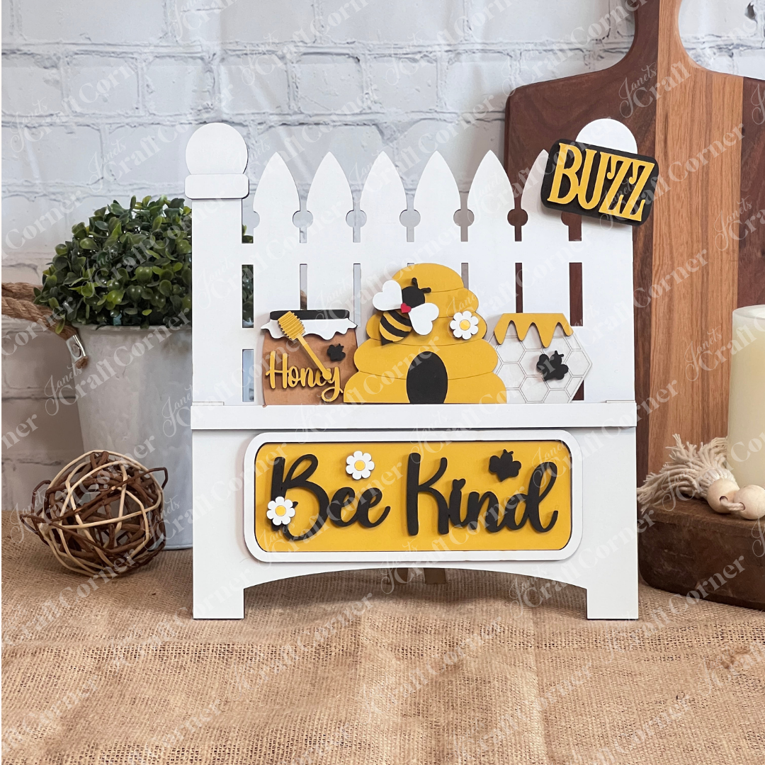 A decorative sign featuring the Bee Interchangeable Insert from Janet's Craft Corner presents a hand-painted white garden fence with a beehive and bees. The piece, which prominently displays "Bee Kind" and a "BUZZ" license plate, stands out against a white brick wall.