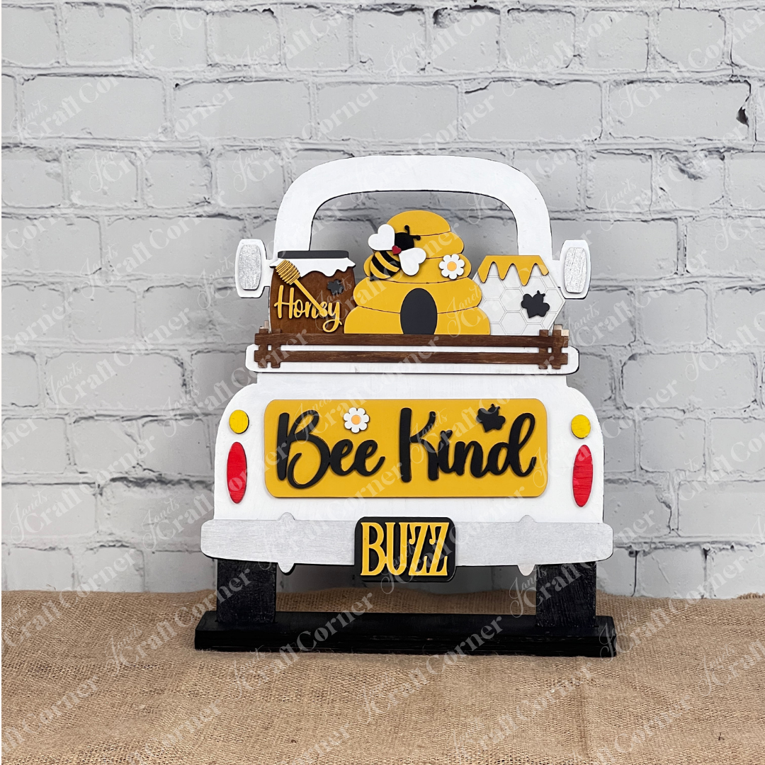 Displayed against a rustic brick wall, Janet's Craft Corner's "Bee Kind Interchangeable Insert" kit showcases a bee-themed truck with a beehive, honey jar, buzzing bees, and the vibrant words "Bee Kind" and "Buzz," all resting on burlap.