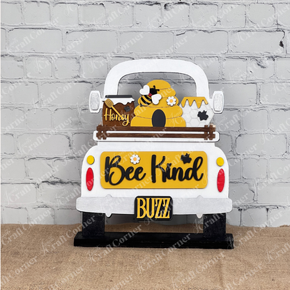 Displayed against a rustic brick wall, Janet's Craft Corner's "Bee Kind Interchangeable Insert" kit showcases a bee-themed truck with a beehive, honey jar, buzzing bees, and the vibrant words "Bee Kind" and "Buzz," all resting on burlap.