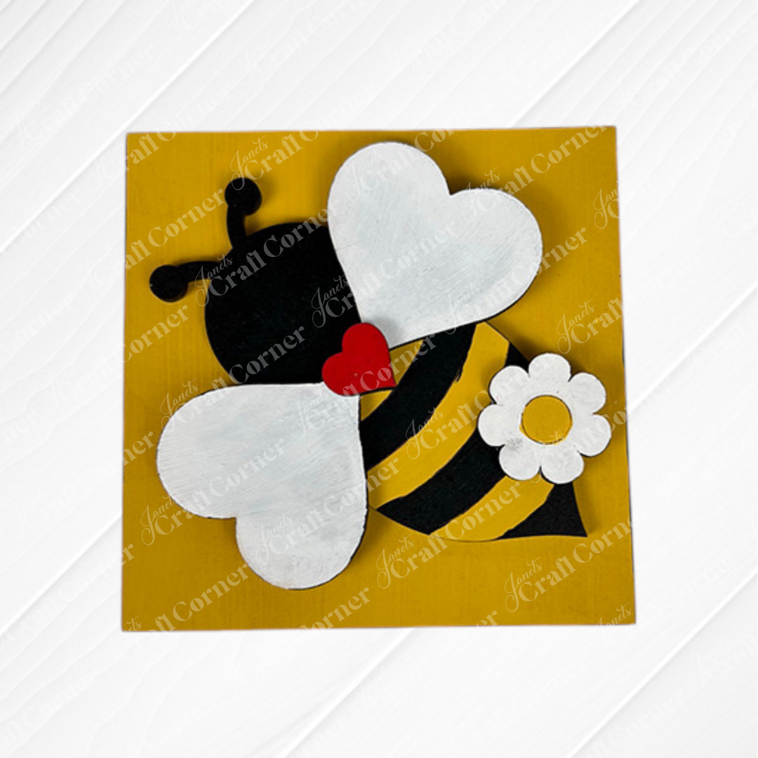 Janet's Craft Corner Bee Swappable Squares feature a handmade bee charm with white wings, black-and-yellow stripes, a red heart, and a white flower on a yellow square—perfect whimsical home decor for any textured white surface.